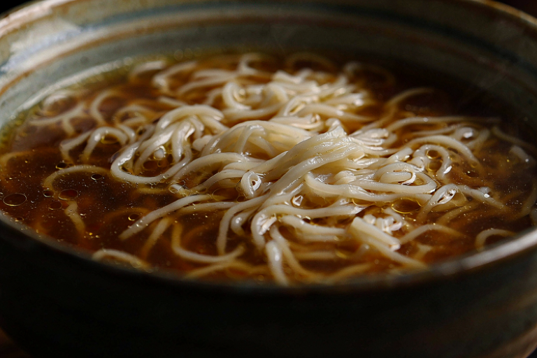 A close-up editorial illustration of a bowl of noodles in a rich broth, emphasizing texture and flavor harmony in a sophisticated dining setting.