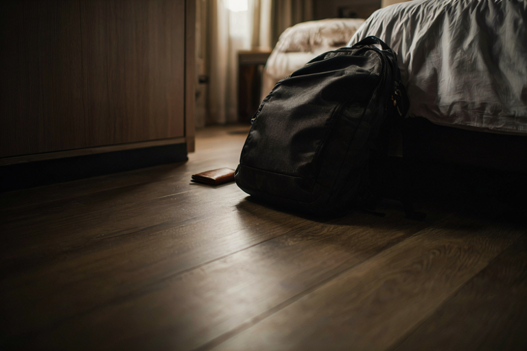 A dark backpack and a leather wallet rest on a polished wooden floor inside a dimly lit hotel room, emphasizing a somber and mysterious atmosphere.