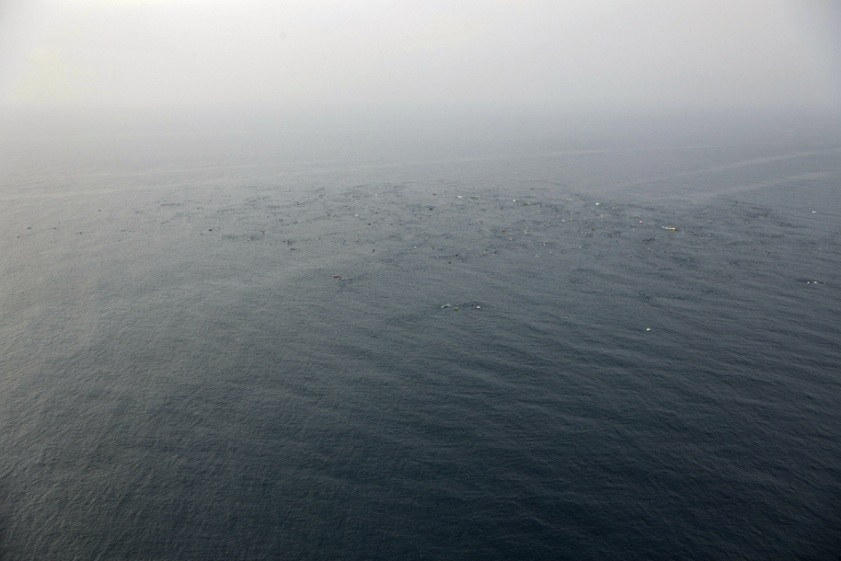 A vast, somber ocean scene off a coastline with a scattered field of debris floating on the water, illustrating an ongoing search operation.