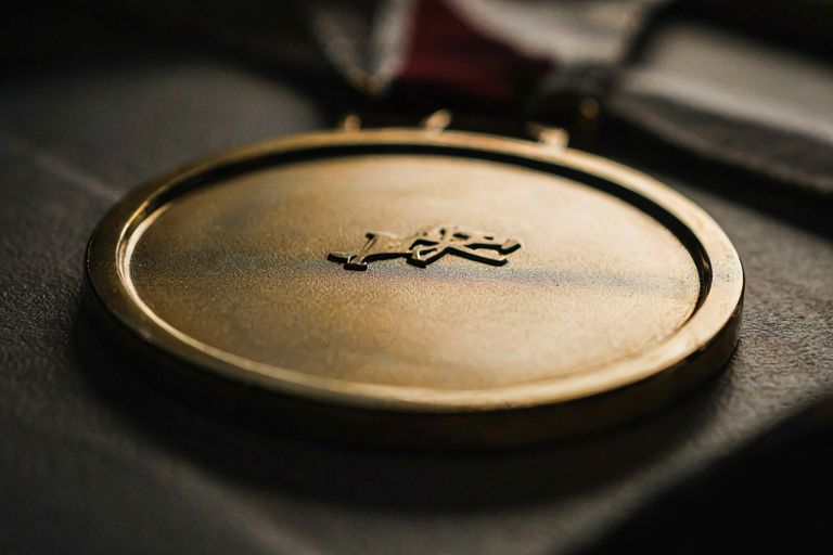 A close-up editorial illustration of a single, polished gold medal resting on a dark, textured surface under focused, dramatic lighting.