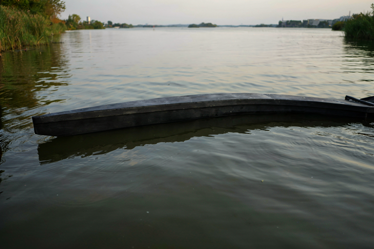 A still, dark body of water reflecting an overcast sky, with the hull of a high-powered model boat resting partially submerged in the foreground.