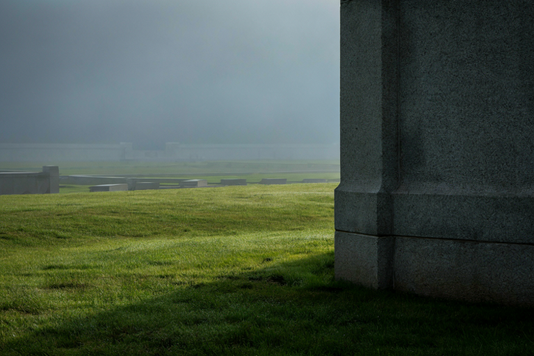 A wide, solemn editorial illustration of a military cemetery at dawn, focusing on a large stone monument amidst low-lying mist and structured rows of markers.