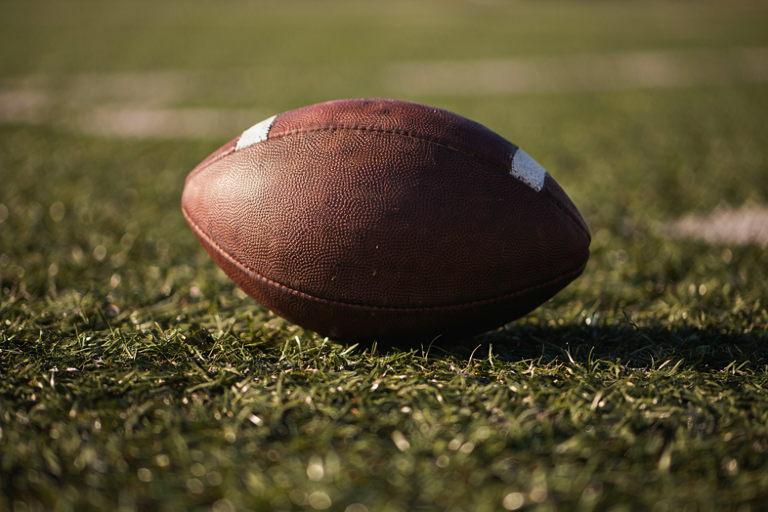 A close-up, dramatic illustration of an American football resting on textured turf, emphasizing the intensity of a high school football game.