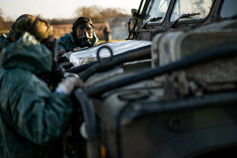 A military vehicle with an Army team in hazmat suits examining it, surrounded by a containment barrier in a rural setting.