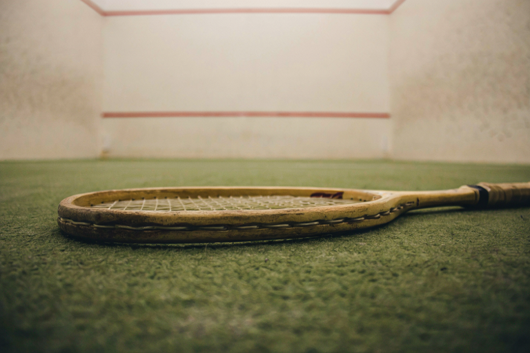 A squash racket on a faded green carpet with a blurred squash court in the background.