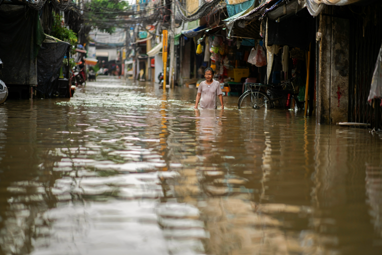A flooded street in Bangkok with a pedestrian wading through knee-deep water, a partially submerged motorbike taxi, and a small grocery store in the background.