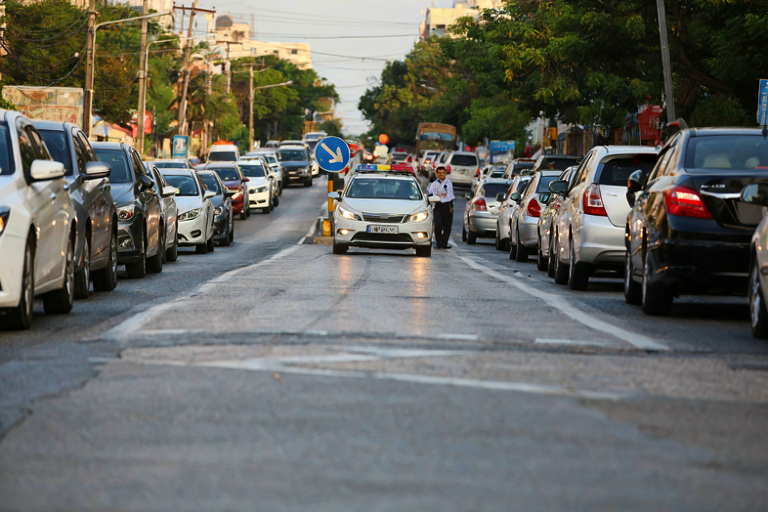 A stalled car on a busy East Bank road with a distant police officer, highlighting traffic issues and potential police presence.