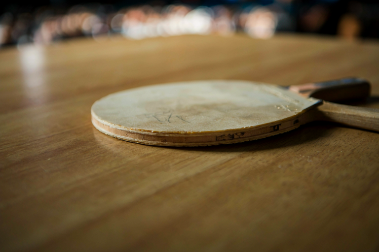 A table tennis paddle resting on a wooden table in a gymnasium, with a blurred crowd in the background.