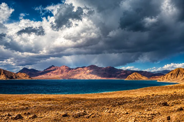 AI illustration based on reference photographs; not an event photograph. A vast, arid mountain landscape featuring a large body of dark blue water bordered by rolling, brown, and reddish mountains under a dramatic, cloudy sky.