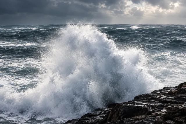 AI illustration based on reference photographs; not an event photograph. A large, powerful wave crashes against a dark, rocky shoreline, generating significant white spray and foam.