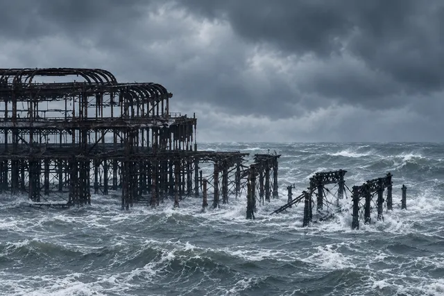 AI illustration based on reference photographs; not an event photograph. A dark, skeletal iron pier structure partially submerged and collapsing into a rough, grey, stormy sea.