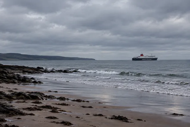 AI illustration based on reference photographs; not an event photograph. A desolate beach scene with a large ferry visible on the water under an overcast sky.