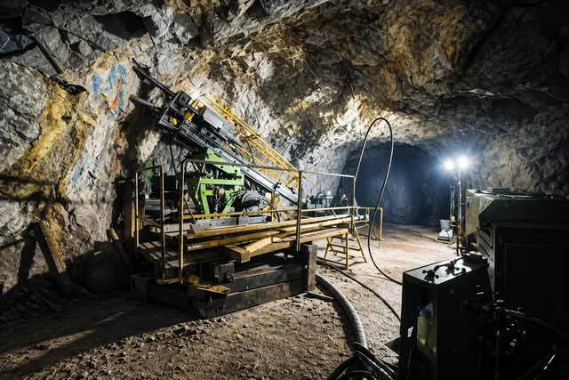 AI illustration based on reference photographs; not an event photograph. A large industrial drilling rig with a complex metal structure is situated inside a rough, rocky cavern with uneven, textured walls.