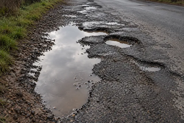 AI illustration based on reference photographs; not an event photograph. Close-up view of a road surface showing numerous large, irregular potholes filled with water and mud.
