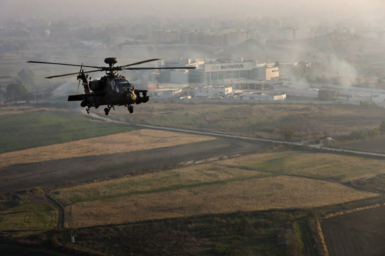 Apache helicopter flying low over a rural landscape with a commercial center in the distance.