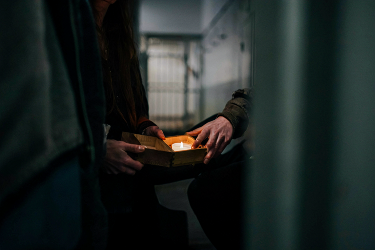 A woman holds a candle in a dim corridor, with a faint prison cell outline visible behind her.