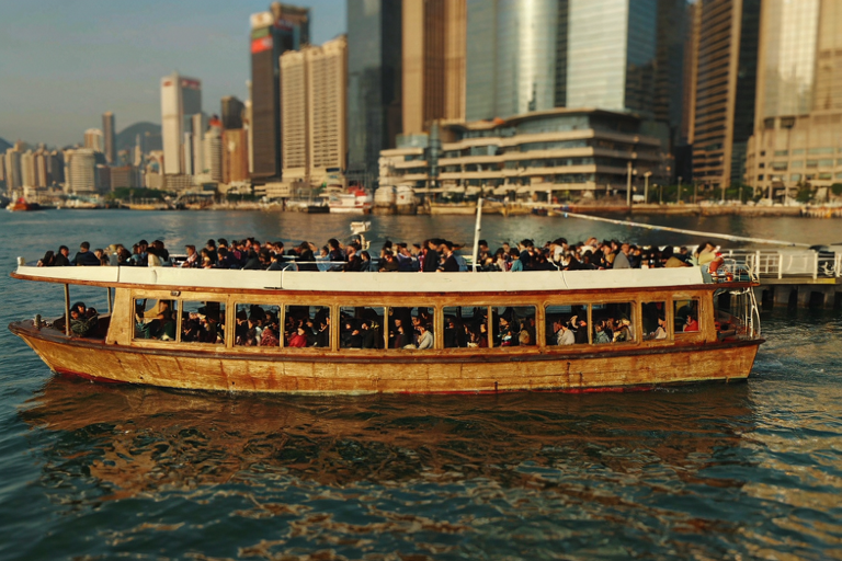 A ferry with passengers in a bustling Hong Kong harbor at dusk, with the city skyline in the background.