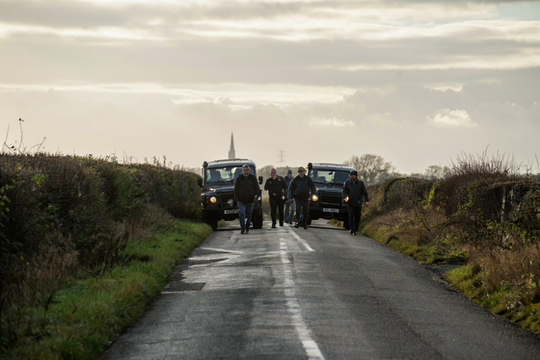 Men blocked by Land Rovers on a narrow road near a church