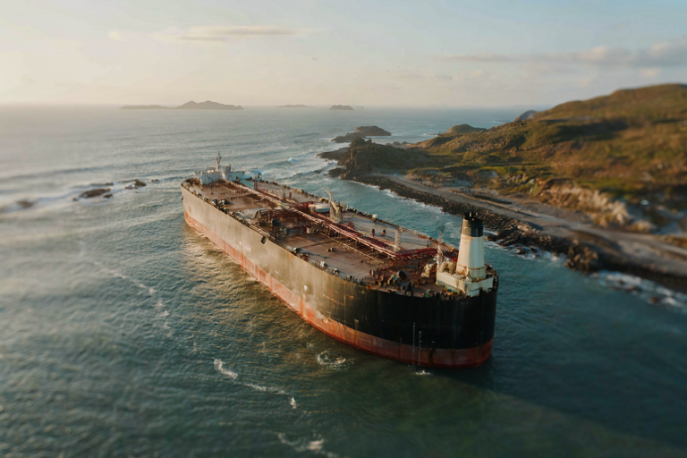 A weathered oil tanker drifts through a narrow strait under golden light.