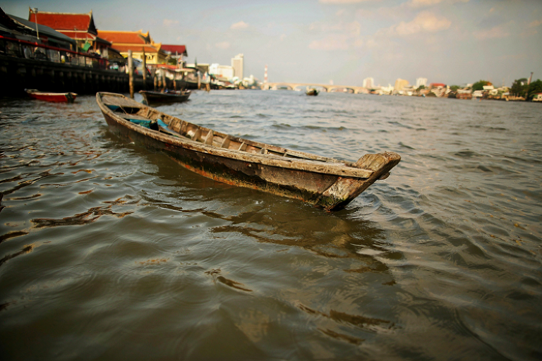 A weathered wooden boat floats in murky water, with distant buildings and a bridge visible through the haze.