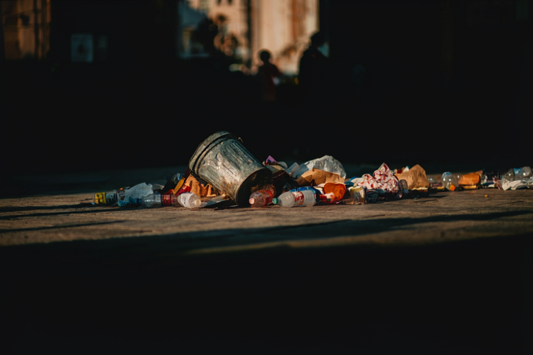 A metal trash can surrounded by litter on a city street at dusk, with blurred figures in the background.