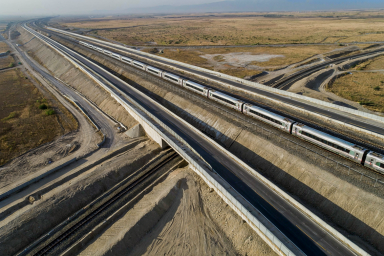 Editorial illustration of a modern high-speed train on an expansive, engineered rail network cutting through a vast landscape.