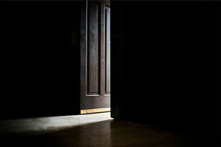 A conceptual illustration of a heavy, dark wooden door slightly ajar, illuminated by a single beam of light, symbolizing an ongoing investigation into a serious campus incident.