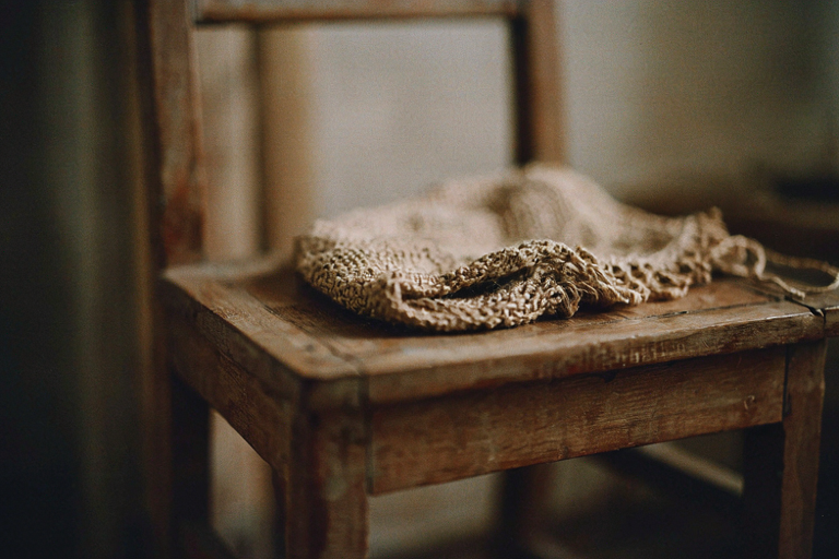 An intimate editorial illustration focusing on a single, aged wooden chair with a woven textile resting upon it, set in a softly lit, warm domestic interior.